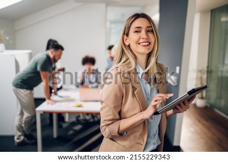 Photo of Smiling confident business leader looking at camera and standing in an office at team meeting. Portrait of confident businesswoman with colleagues in boardroom. Using digital tablet during a meeting.