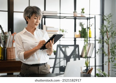 Smiling confident business leader standing in an office. Mature asian business woman wearing white shirt using tablet standing in cozy workspace. - Powered by Shutterstock