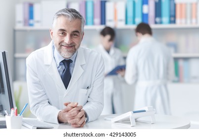 Smiling confident doctor at the reception desk, medical staff working on the background - Powered by Shutterstock