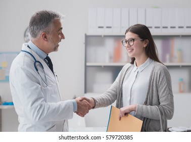 Smiling confident doctor shaking hands with a female patient in the office, she is holding medical records - Powered by Shutterstock