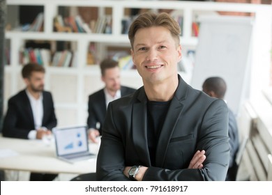 Smiling confident executive in suit standing with arms crossed looking at camera with business partners team at background, happy company ceo co-founder boss board member headshot portrait