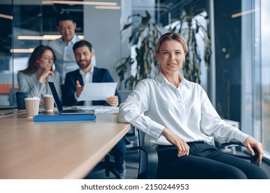 Smiling Confident Female Boss Sitting On Meeting In Office With Her Colleagues At Background. Business Concept