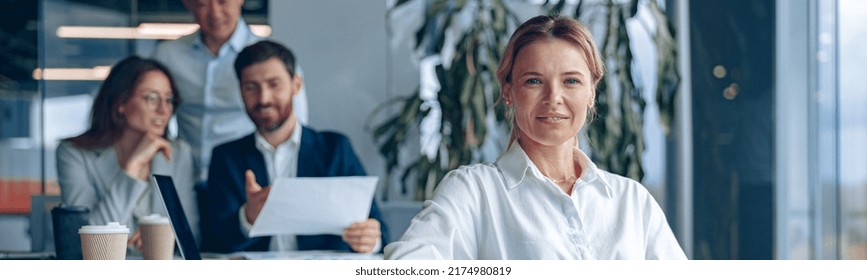 Smiling Confident Female Boss Sitting On Meeting In Office With Her Colleagues At Background.