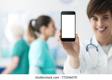 Smiling Confident Female Doctor Holding A Touch Screen Smartphone And Medical Staff On The Background, Medical App Concept