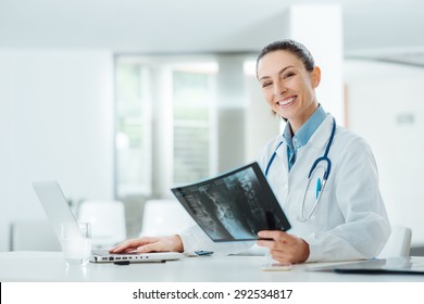 Smiling confident female doctor sitting at office desk and examining a patient's x-ray, she is looking at camera - Powered by Shutterstock