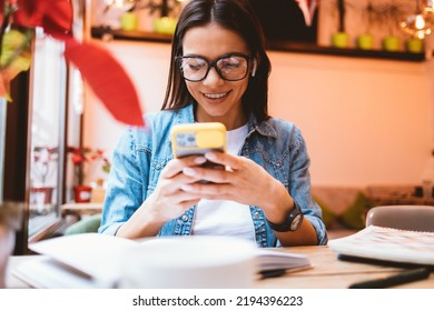 Smiling Confident Female Student Using Her Smartphone Sitting At Table With Notebooks And Textbooks