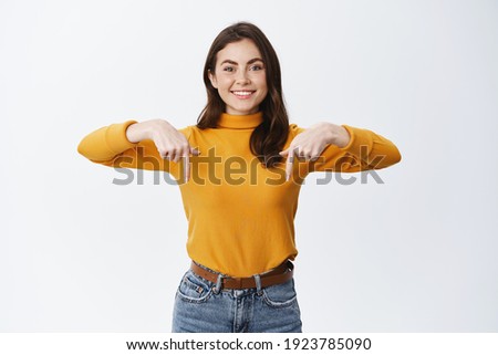Smiling confident girl pointing fingers down to show advertisement, showing promo offer on bottom empty space, standing against white background.