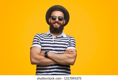 Smiling confident hipster guy in hat and sunglasses holding arms crossed looking at camera on yellow background