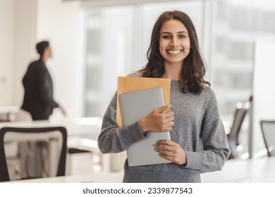 Smiling confident Indian woman wearing casual clothes holding laptop and documents, looking at camera at modern office. Good looking businesswoman posing for picture. Concept of successful business 