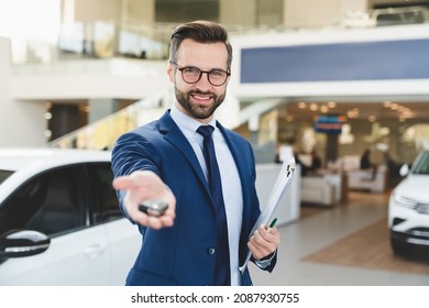Smiling confident male shop assistant in formal clothes holding handing car keys to a client after buying purchasing new expensive car at automobile dealer shop.