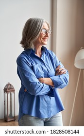 Smiling Confident Mature Woman Standing Arms Crossed Indoors Looking At Window. Stylish Elegant Middle Aged Senior 60s Gray-haired Lady Wearing Glasses Thinking Of Future Vision, Dreaming At Home.