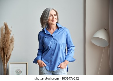 Smiling Confident Mature Woman Standing Indoors Looking At Window. Stylish Elegant Middle Aged Senior 60s Gray-haired Lady Thinking Of Good Future Vision, Enjoying Wellbeing, Dreaming At Home.