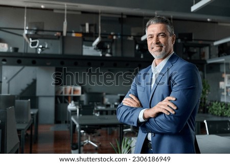 Smiling confident middle aged business man, mature older professional successful company ceo corporate leader wearing blue suit standing in modern office with arms crossed looking at camera, portrait.