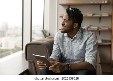 Smiling Confident Prosperous African Male Employee Take Break Sit On Sofa In Modern Office Room Holding Digital Tablet Looking Out Panoramic Window. Daydreams, Business Vision And Aspiration Concept