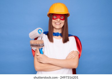 Smiling confident renovation professional woman in superhero costume holding a paintbrush and roller keeps arms crossed ready for help with house repair work isolated over blue background - Powered by Shutterstock