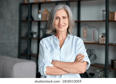 Smiling confident stylish mature middle aged woman standing at home office. Old senior businesswoman, 60s gray-haired lady executive business leader manager looking at camera arms crossed, portrait.