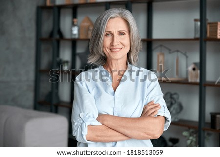 Photo of Smiling confident stylish mature middle aged woman standing at home office. Old senior businesswoman, 60s gray-haired lady executive business leader manager looking at camera arms crossed, portrait.