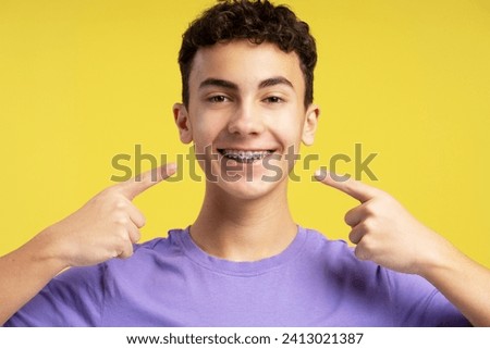Smiling confident teenager pointing fingers on dental braces looking at camera isolated on yellow background. Health care, hygiene, orthodontic concept