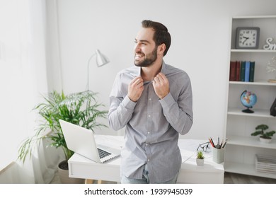 Smiling Confident Young Bearded Business Man In Gray Shirt Stand Near Desk With Laptop Pc Computer In Light Office On White Wall Background. Achievement Business Career Concept. Straightening Collar