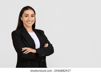 Smiling confident young professional woman with dark hair, wearing black suit and white shirt, standing with arms crossed on light background, copy space - Powered by Shutterstock