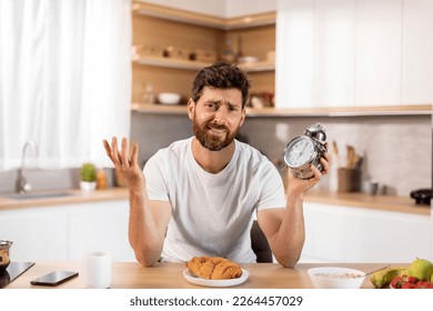 Smiling confused tired sleepy adult european man in t-shirt hold alarm clock, spreads his arms to sides at table in modern kitchen interior. Breakfast, good early morning alone at home, time for work - Powered by Shutterstock