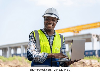 Smiling Construction Engineer Using Laptop on Site, Happy African American Worker with Computer at Project Site, Man in Hard Hat and Vest Working Outdoors