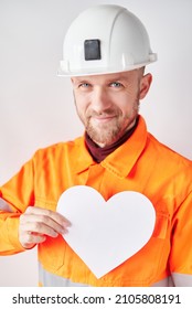 Smiling Construction Worker Or Engineer In Hard Hat And Orange Vest With White Paper Heart As Valentine's Day Symbol. Construction Industry Or Romance Concept. High Quality Image