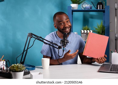 Smiling Content Creator Reviewing Red Cover Mockup Book Talking Using Professional Microphone Sitting At Desk With Laptop. Influencer Presenting A New Novel Volume On Social Media.