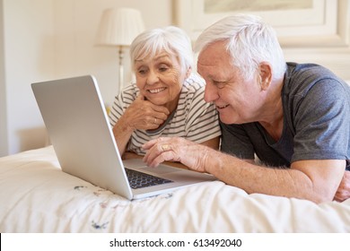 Smiling and content senior couple talking and using a laptop while lying in bed together in the morning - Powered by Shutterstock