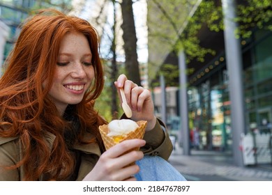 Smiling Cool Teenage Hipster Girl Eating Ice Cream On Big City Urban Street. Pretty Teen Generation Z Girl With Red Hair Enjoying Delicious Tasty Icecream Spending Time In Park Outdoors.