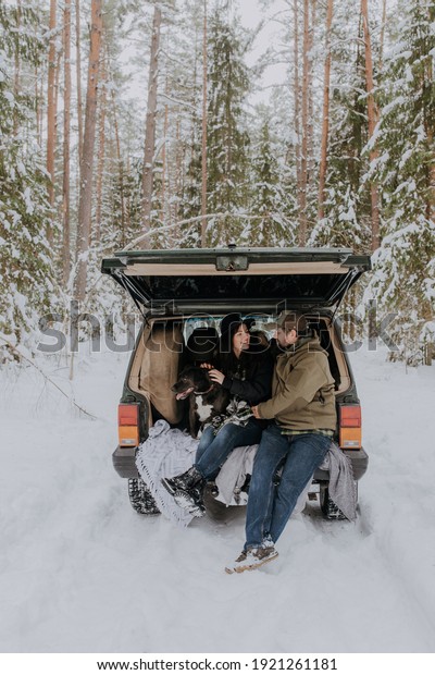 Smiling couple with black dog sitting\
in open SUV car trunk in snowy forest. Enjoying each other in\
active winter holidays. Active lifestyle\
concept.