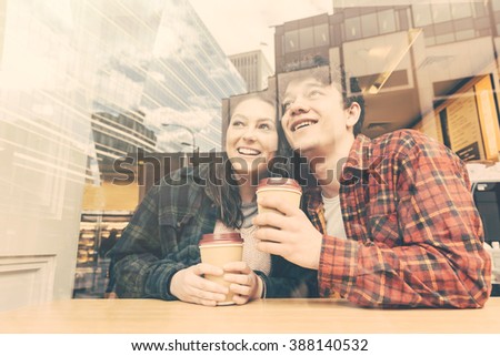 Smiling couple in a cafe in London. They are a boy and a girl, holding a cup of tea or coffee and looking out of the window with an happy face. Photo taken from outside, seen through the glass window