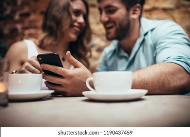 Smiling Couple At A Coffee Shop Spending Time Together Talking. Man Sitting At A Restaurant With His Girlfriend And Looking At His Mobile Phone.