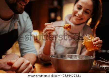 Smiling couple in a cozy kitchen adding honey to a mixing bowl. They are enjoying baking, creating a warm and inviting atmosphere with natural ingredients and teamwork
