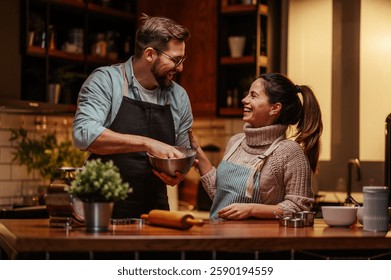 Smiling couple in a cozy kitchen preparing a meal. They are wearing aprons and sharing a joyful moment while mixing ingredients. The warm lighting adds a homely atmosphere to the scene