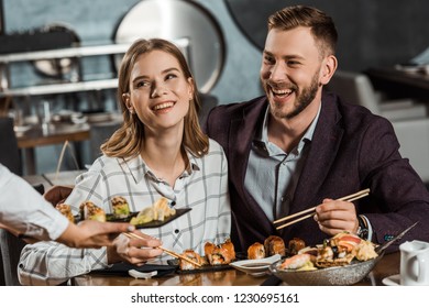 Smiling Couple Eating Sushi Rolls While Waiter Bringing New Order In Restaurant