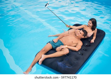 Smiling Couple Floating On Mattress In Pool And Taking Selfie Photo On The Phone With Selfie Stick On Their Summer Vacation. Copy Space