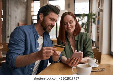 Smiling couple of friends holding credit card, using mobile phone shopping online, drinking coffee  sitting in modern cafe. Successful freelancers receive payment                                - Powered by Shutterstock