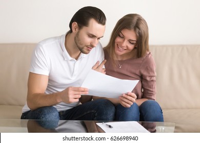 Smiling Couple Holding Papers And Carefully Studying Documents Sitting On Sofa Indoors, Reading Terms And Conditions, Reviewing Agreement, Considering Mortgage Loan Offer, Personal Insurance 