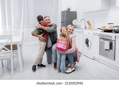 Smiling Couple Hugging Grandchildren With Backpacks In Kitchen