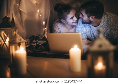 Smiling Couple Lying On Bed Looking At Each Other With A Laptop In Front. Couple Enjoying A Movie On Laptop At Night In Bedroom Lit With Candles And Tiny Bulbs.