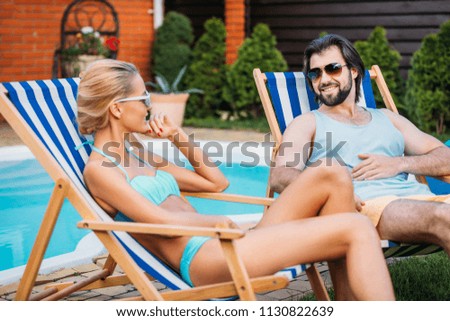 smiling couple on beach chairs spending time near swimming pool on backyard on summer day