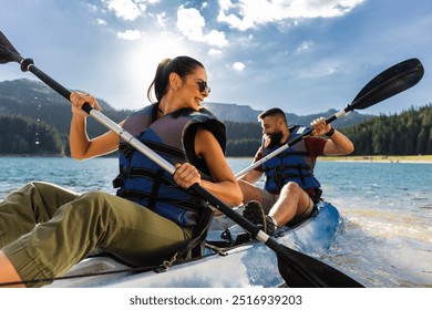 A smiling couple paddles together in a kayak on a scenic lake, surrounded by mountains and clear skies.