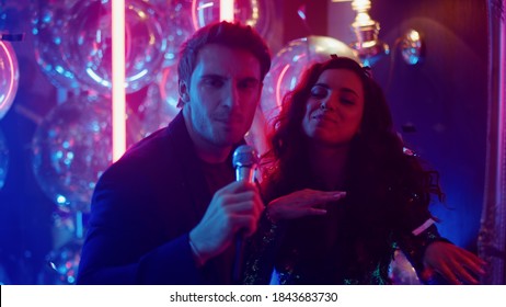 Smiling Couple Performing Song At Party In Night Club. Young Man And Woman Singing At Karaoke On Neon Lamps Background. Handsome Man Holding Microphone In Nightclub.