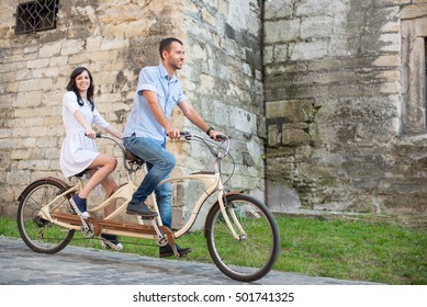 Smiling couple riding on retro tandem bicycle against the background of the old brick building. Man looks forward and the girl in a white dress looking at the camera. Lviv, Ukraine - Powered by Shutterstock