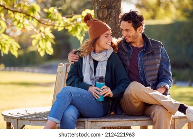 Smiling Couple Sitting On Bench In Autumn Park With Reusable Drinks Cups