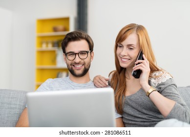 Smiling Couple Sitting On Sofa With Laptop Computer And Cordless Telephone, Placing An Order Or Shopping Online