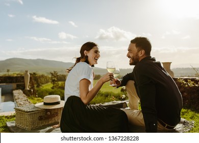 Smiling Couple Sitting In A Wine Farm Holding Glasses Of White Wine. Couple On A Date Sitting In A Vineyard With A Picnic Basket By Their Side.