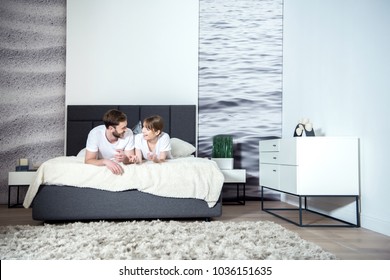 Smiling Couple Talking And Lying On Bed In Cozy Bedroom With Modern Interior