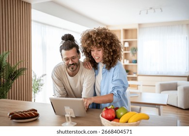 Smiling couple using digital tablet, browsing internet or watching movie, enjoying leisure time at home, healthy lifestyle with fresh fruits and bread in modern kitchen